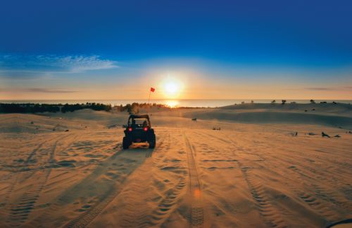 The image is of a clear blue sky with the sun setting below with an orange haze. There is a dome buggy with a red flag on top driving through the soft sand at the Silver Lake Sand Dunes.