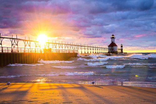 The image is of a cloudy sunset at St. Joseph Lighthouse. White cascading waves are flowing towards the lighthouse at the end of the pier. 