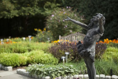 The image is of a garden in the background with flowers and bushes. In focus, there is a statue of a little girl.