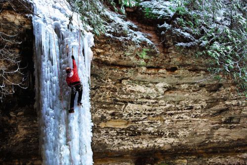 The image is of an ice climber to the left in a red jacket with ice picks dug into sheets of ice. To the right is a rocky surface draped with trees in a light frost of snow.