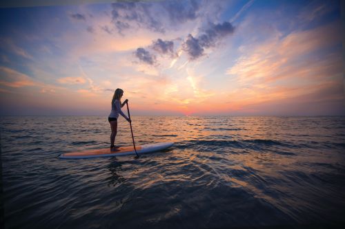 The image is a beautiful orange, blue and purple sunset with an endless lake of water. There is a woman silhouette on a paddleboard paddling in Grand Haven.