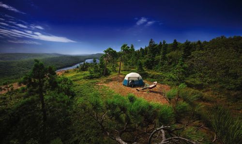 The image of the beautiful Porcupine Mountains. A camper near his tent is surrounded by beautiful forest green trees. With a big blue sky with clouds floating by.