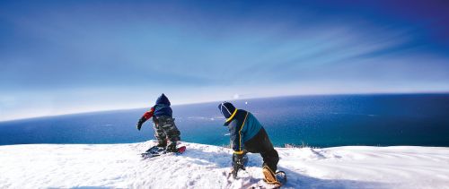 The image is Winter at Sleeping Bear Dunes. There are two people snowshoeing looking over a beautiful dark and light blue Lake. 
