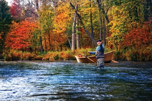 The image is a beautiful fall day in Pere Marquette. There is blue and green water of a lake with a man in a hat and glasses, fly fishing. Next to the man is a small wooden boat with paddles. 
