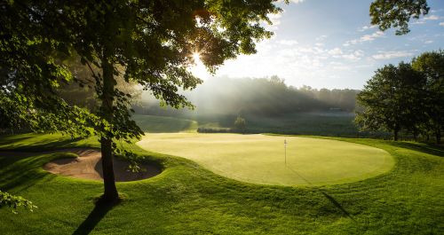 The image is of a blue cloudy sky with the sun peaking through a full tree. The sun is casting a haze over the beautiful bright green golf course with a sign flag at Pilgrims Run.